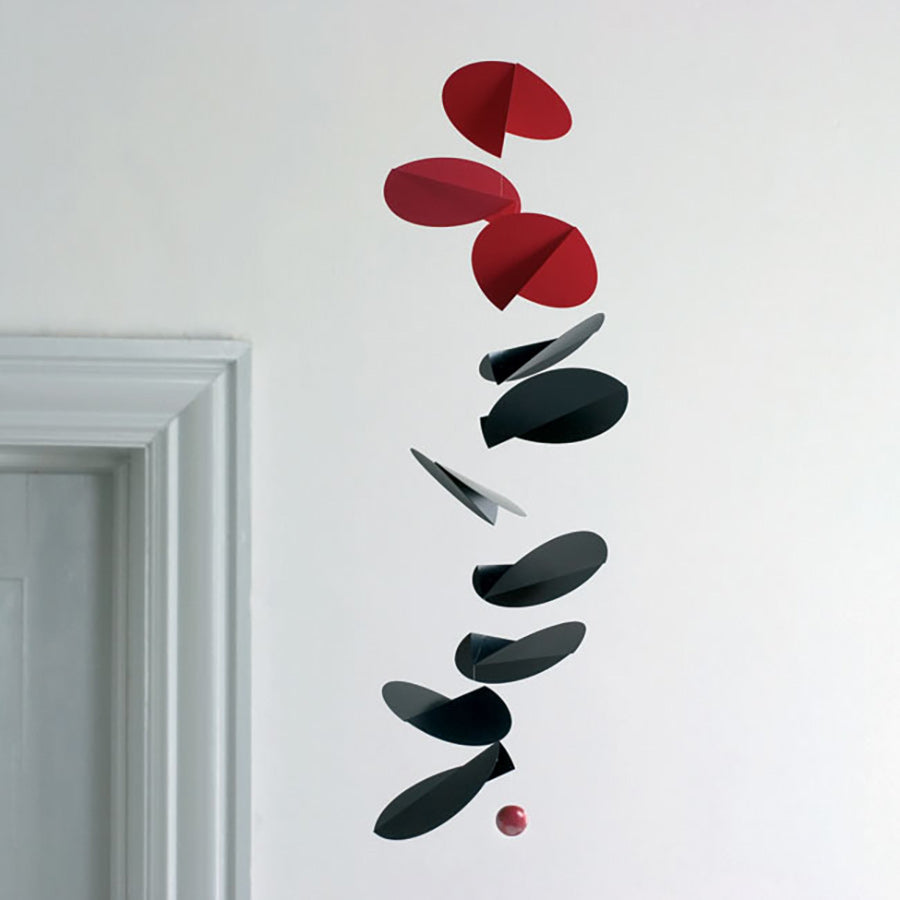 The Turning Leaves Mobile by Flensted features abstract, flat circular shapes in red, black, and gray, hanging gracefully from the ceiling near a white doorframe and creating a modern artistic display against a plain wall.