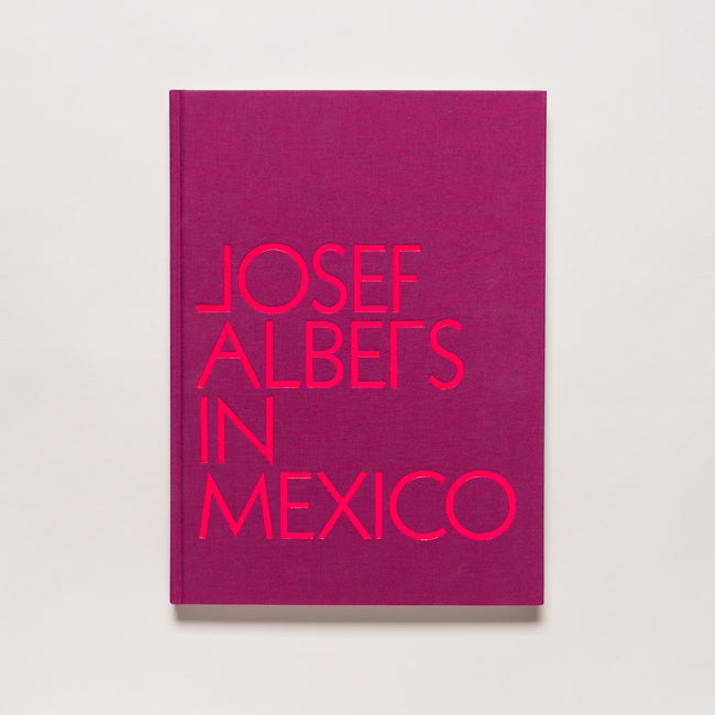 The book "Josef Albers in Mexico" features bold, bright pink text on a magenta cover and is photographed against a plain white background, highlighting abstract art influenced by pre-Columbian architecture.