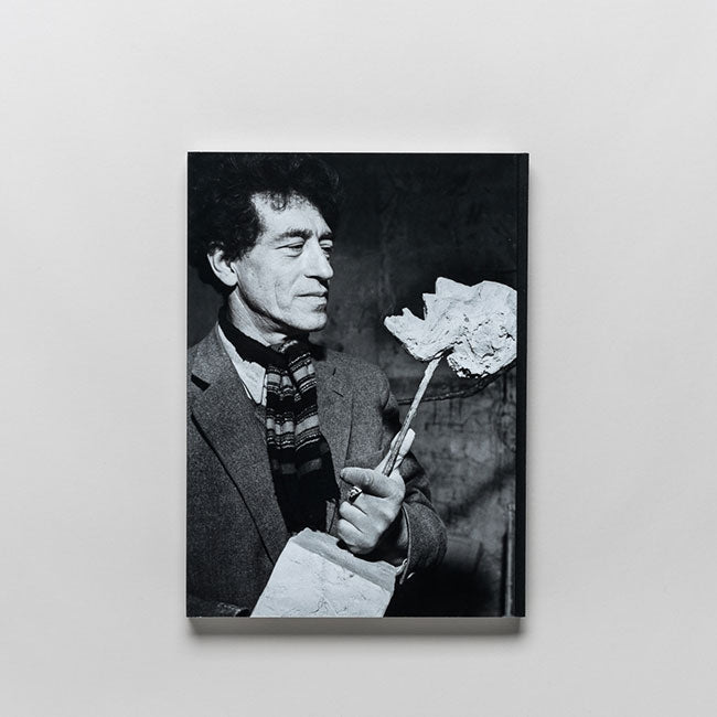 Black and white photo of a man in a suit and striped scarf holding a sculpted face on a stick, evoking the iconic figurative style of Giacometti.