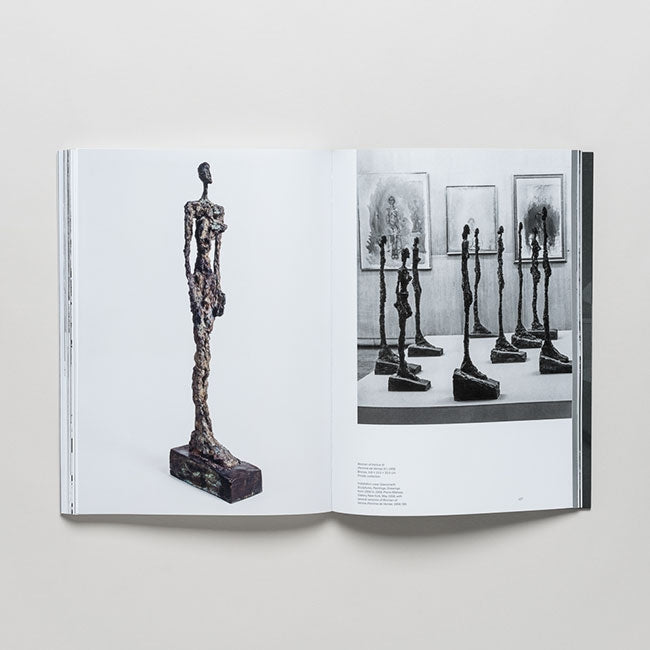 An open art book titled "Giacometti" displays a tall, slender bronze sculpture by Alberto Giacometti on the left page and a group of similar figures in a gallery setting on the right.