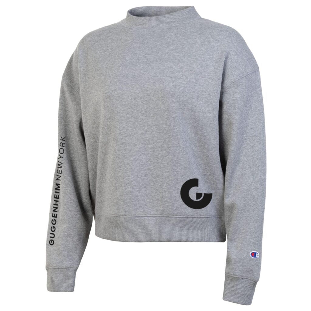 The Guggenheim Women's Triumph Tri Blend Crew Sweatshirt is heather gray with long sleeves, a soft poly-cotton blend, GUGGENHEIM NEW YORK on the left sleeve, a black G logo front, and a Champion logo on the left wrist.