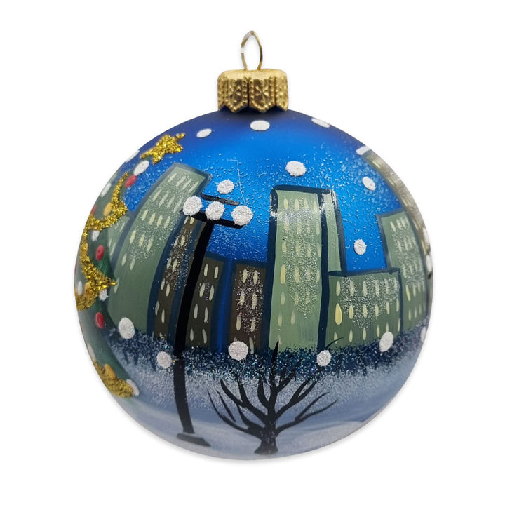 The NYC Tree Ornament is a round blue glass ornament, mouth-blown and hand-painted with a cityscape of tall green buildings, snowy ground, and trees. Red dots and gold glitter decorate one side like a festive tree. Comes gift-boxed.