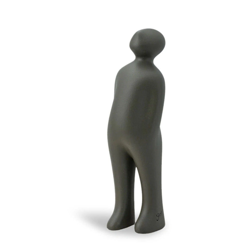 The Visitor Sculpture, Small (Graphite) is a minimalist dark gray ceramic figurine inspired by Guido Deleu, depicting a standing human figure with a rounded head and simplified body against a white background.