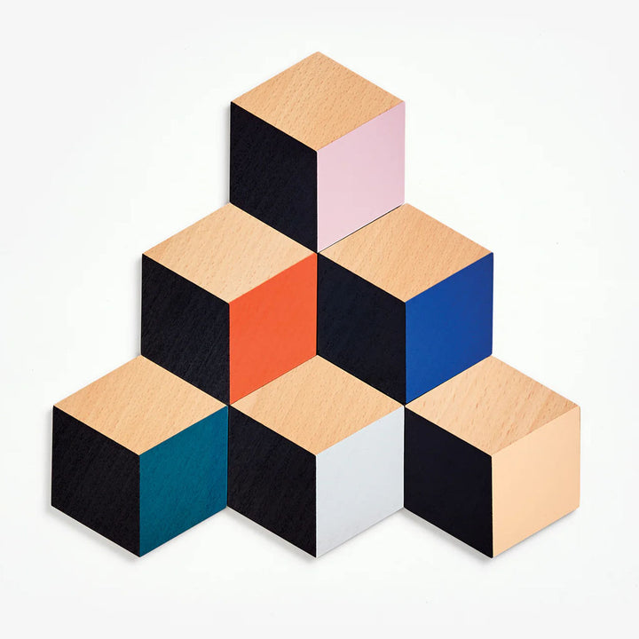The Table Tiles Geometric Trivet/Coaster Set features seven wooden cubes with faces in black, wood, pink, orange, blue, teal, and white—arranged in a modern geometric pattern and perfect as a stylish housewarming gift.