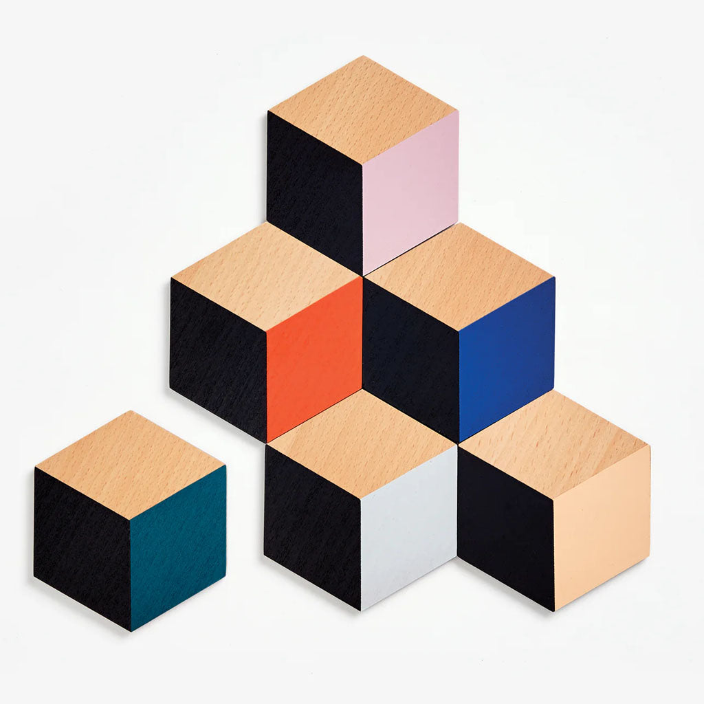The Table Tiles Geometric Trivet/Coaster Set features seven wooden cubes in black, natural wood, pink, orange, blue, teal, and white. Arrange them for a modern look—perfect as unique coasters or a special housewarming gift.