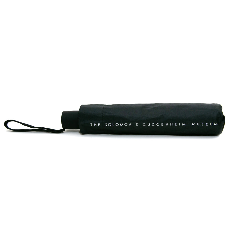 The Guggenheim Skylight Umbrella is a closed black umbrella with a wrist strap, featuring "THE SOLOMON R GUGGENHEIM MUSEUM" in white letters—both a stylish museum gift and practical accessory.
