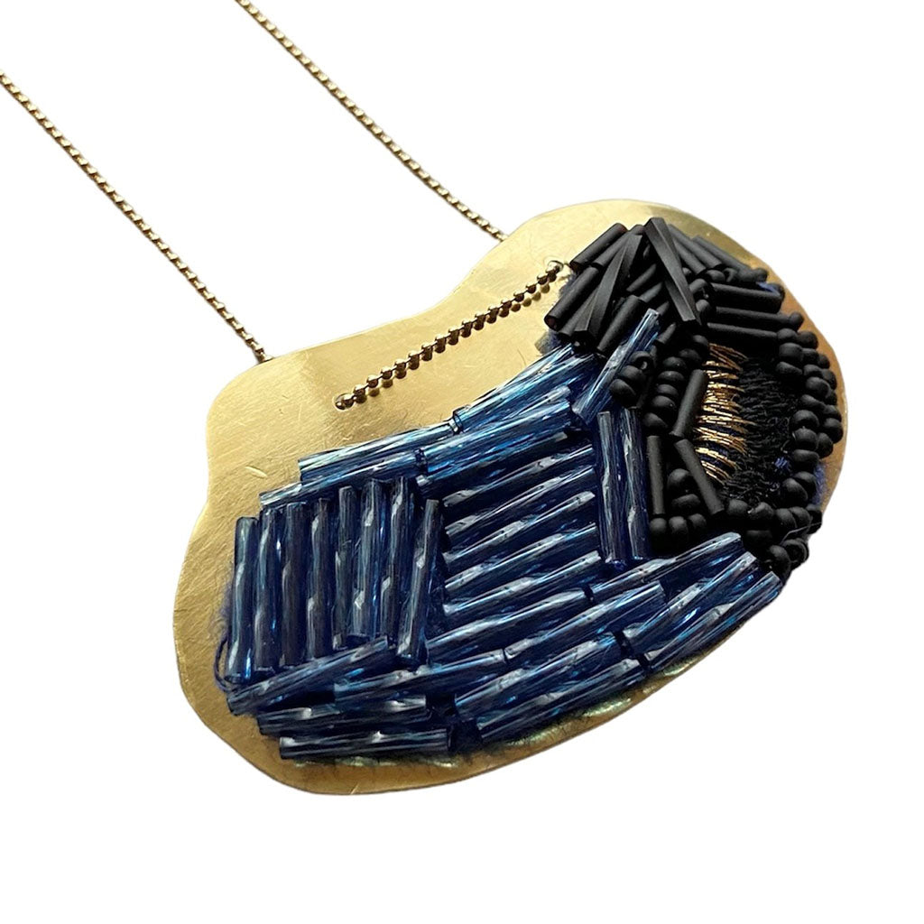The Trecy Bleich Rice Field Necklace Denim/Charcoal features a gold pendant with an abstract design, highlighted by textured blue and black beads arranged in geometric patterns.