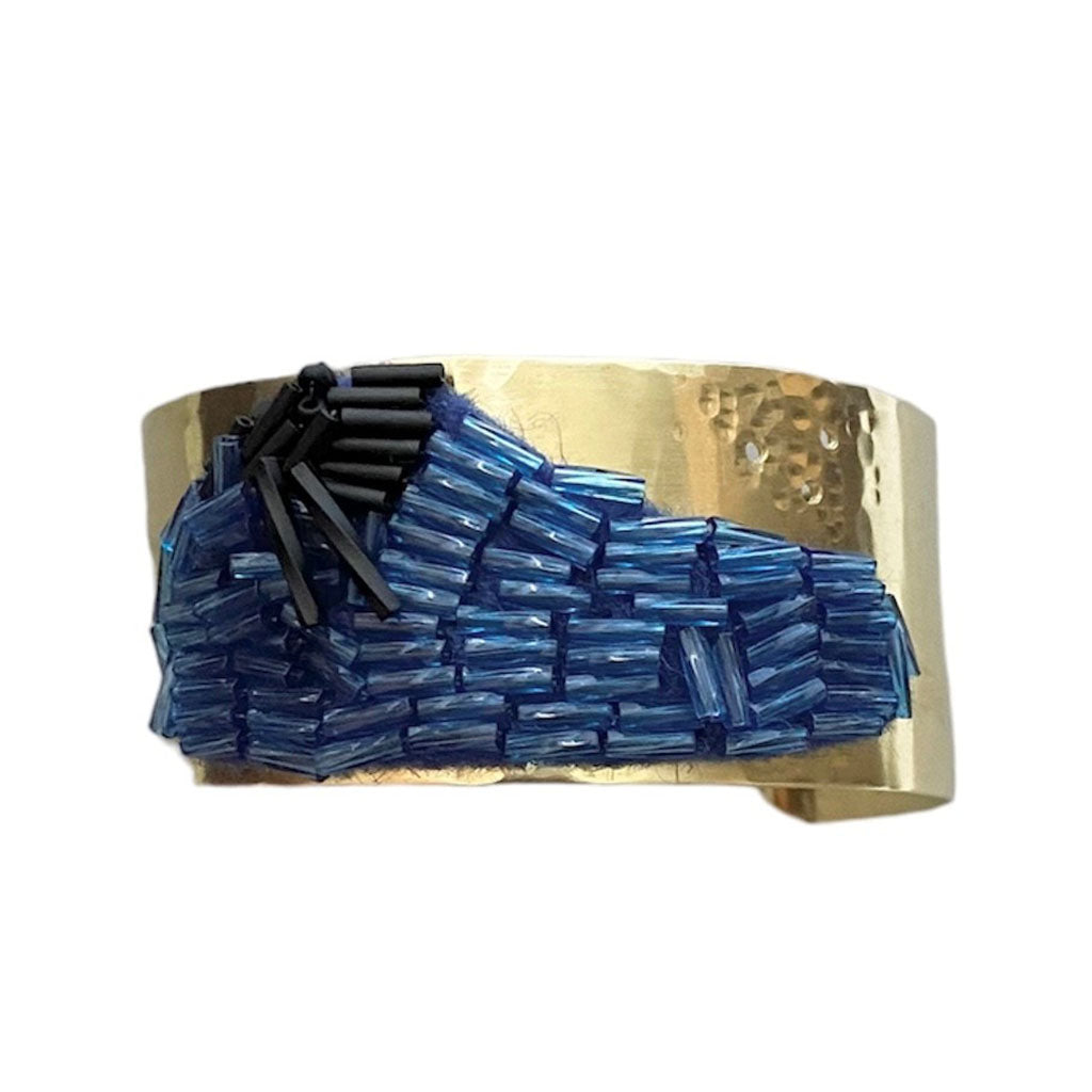 The Trecy Bleich Rice Field Bangle Bracelet in Denim/Charcoal features a wide, hammered gold band with wave patterns of blue and black beads, evoking ocean waves—a stunning handcrafted piece from the Rice collection.