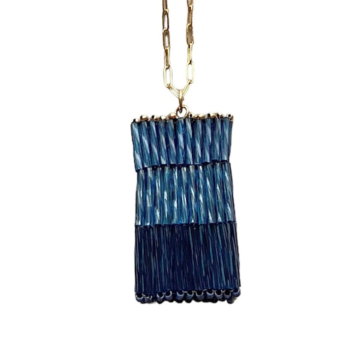 The Trecy Bleich Rice Stack Necklace Denim features a rectangular pendant with layered blue and navy woven segments, suspended from a gold elongated link chain.