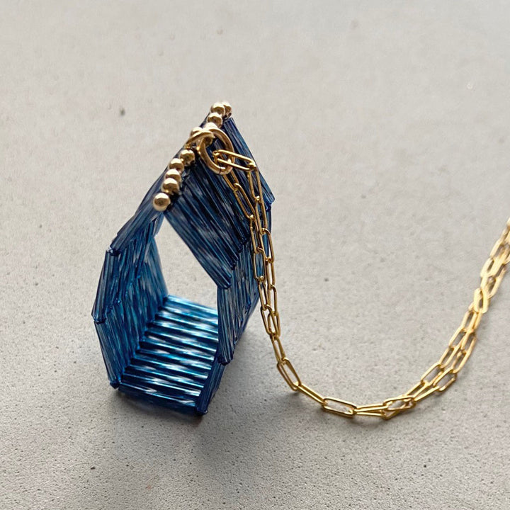 The Trecy Bleich Rice Stack Necklace Denim features a gold chain threaded through a geometric, house-shaped blue pendant of vertically aligned rods with an open center. This handcrafted piece from the Rice collection rests on a light gray surface.