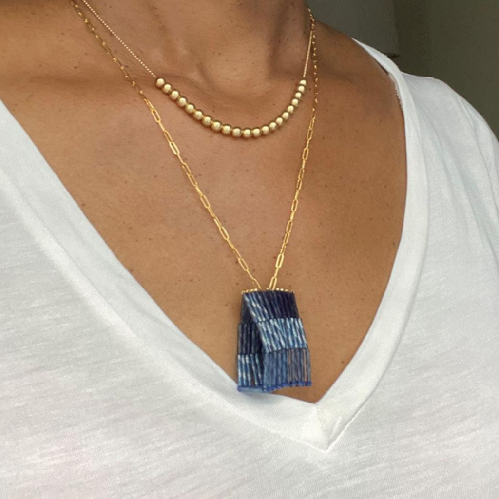 A person wearing a white V-neck shirt, handcrafted jewelry including a gold chain with small pearls, and the Trecy Bleich Rice Stack Necklace Denim—a longer gold necklace with a blue rectangular fringed pendant.