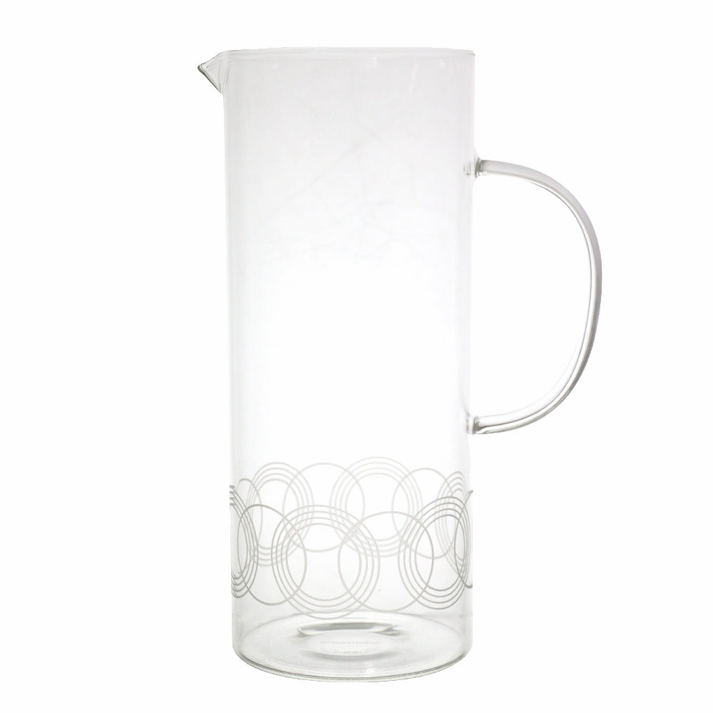 The Guggenheim Gates Glass Pitcher, inspired by Frank Lloyd Wright, features a handle and a subtle pattern of overlapping silver circles near the base. It is shown against a plain white background.