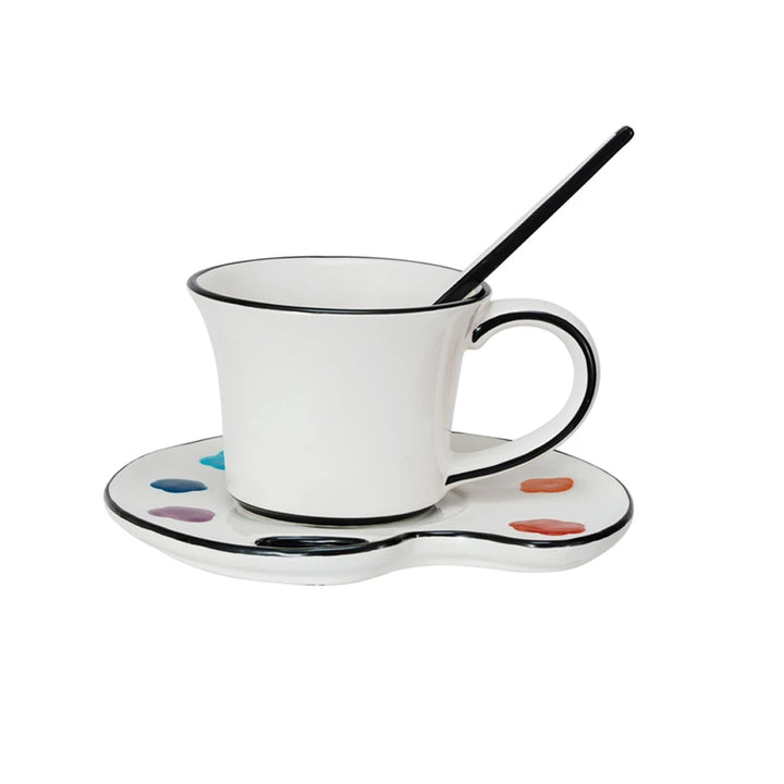 The Ceramic Paint Palette Saucer w/ Teacup & Brush Shaped Stirrer, Set of 3 features a white teacup with a black rim and handle on an artist palette saucer with colorful paint splotches plus a brush-shaped stirrer.