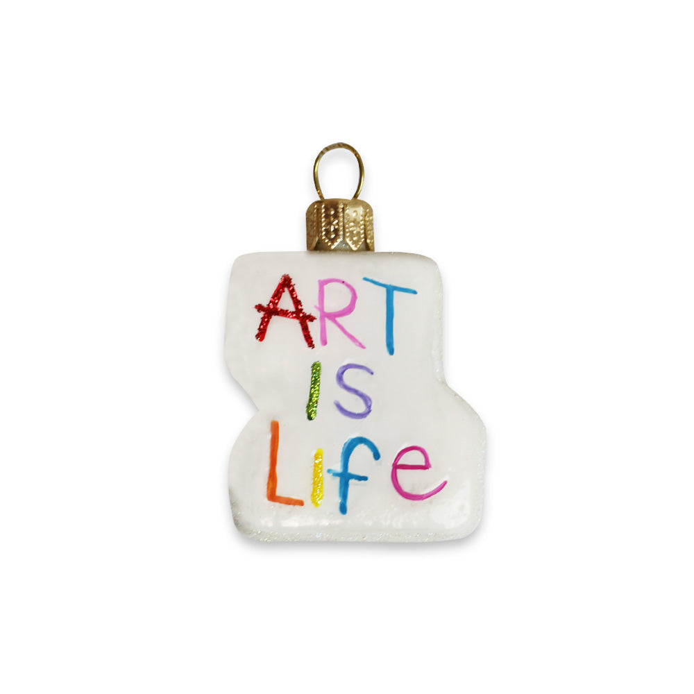 The Art is Life Ornament is a white blown glass paint palette with a gold cap and colorful "ART IS LIFE" lettering on the front, inspired by Humberto Cruz.