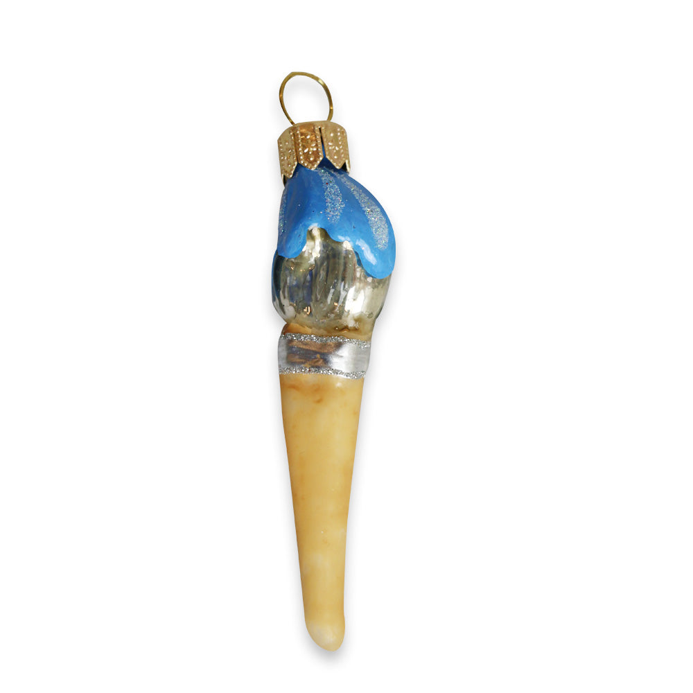 A blown glass ornament shaped like a torch with a gold cap, blue and silver flame-like top, and long beige pointed stem, set against a white background—an Art Icons Mini Paintbrush Ornament.