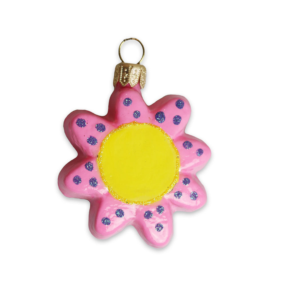 The Art Icons Mini Flower Ornament by Humberto Cruz is a flower-shaped blown glass piece with pink petals, purple dots, a bright yellow center, and a gold cap with hanging loop, shown on a white background.