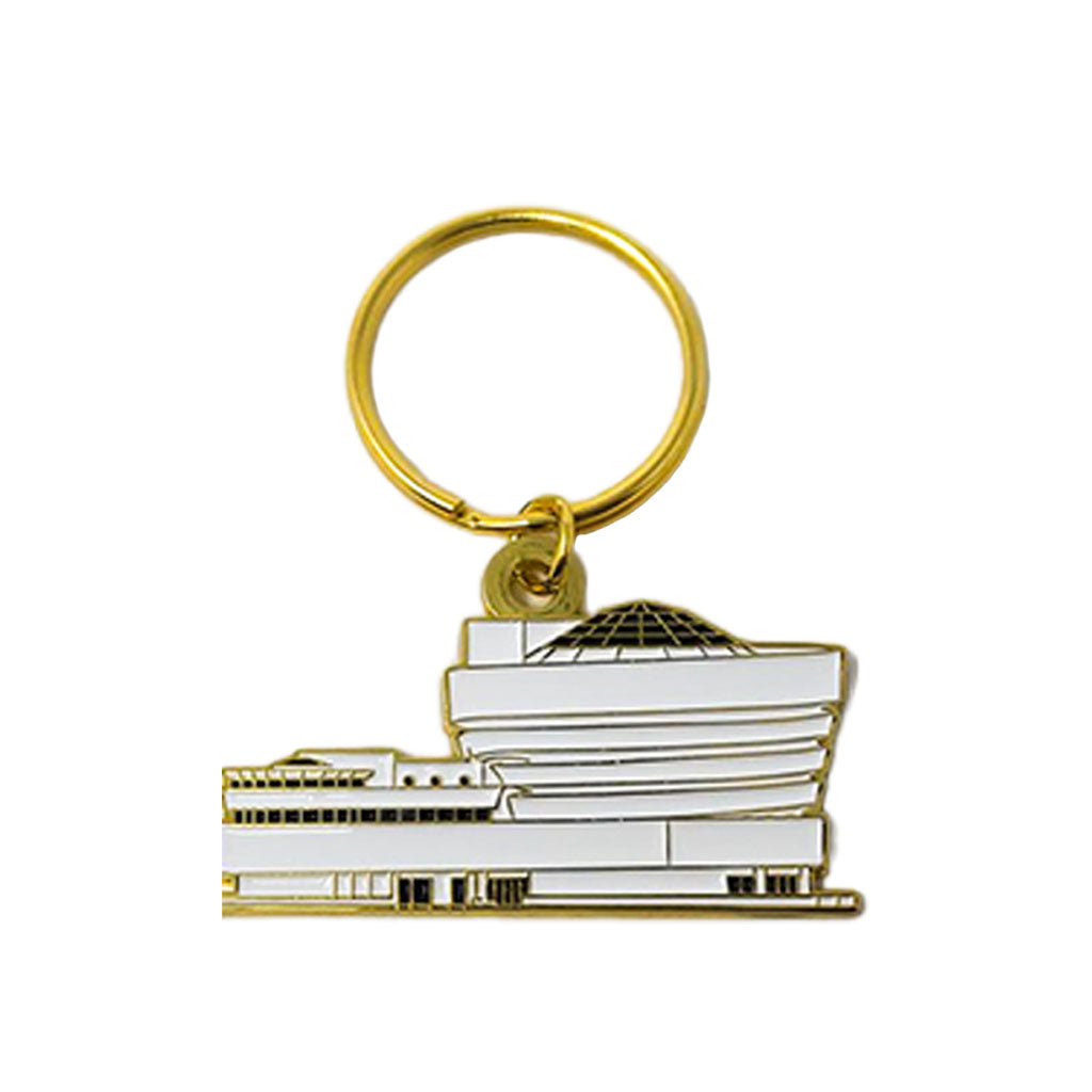 A gold keyring holds the Guggenheim Building Enamel Keychain, shaped like a stylized white building with a glass dome and modern design. The background is plain white.