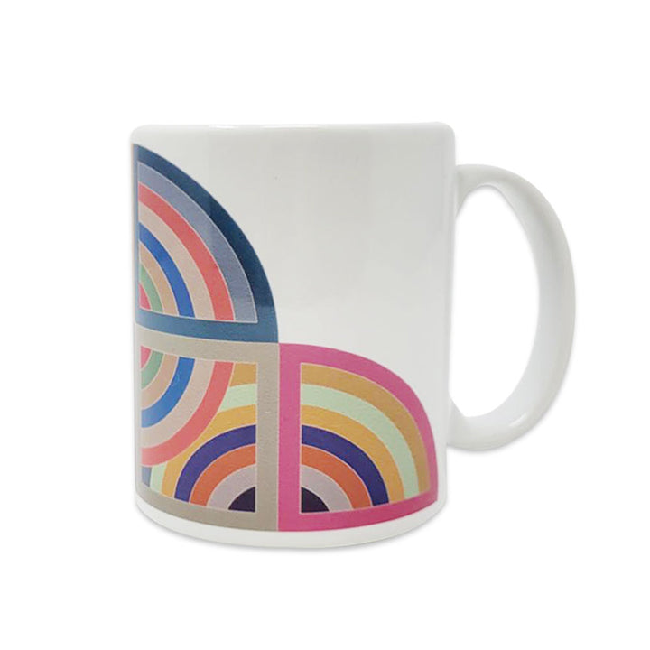 The Stella Harran II Mug is a white ceramic coffee mug with a vibrant geometric design of overlapping pink, blue, yellow, and orange semi-circles and arcs on one side.