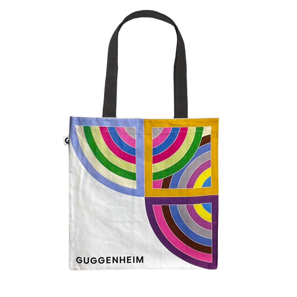The Stella, Harran II Tote is a white cotton tote with black handles and colorful overlapping semicircles in blue, green, pink, red, and yellow. "GUGGENHEIM" appears in black at the lower left—it's a bold statement bag.