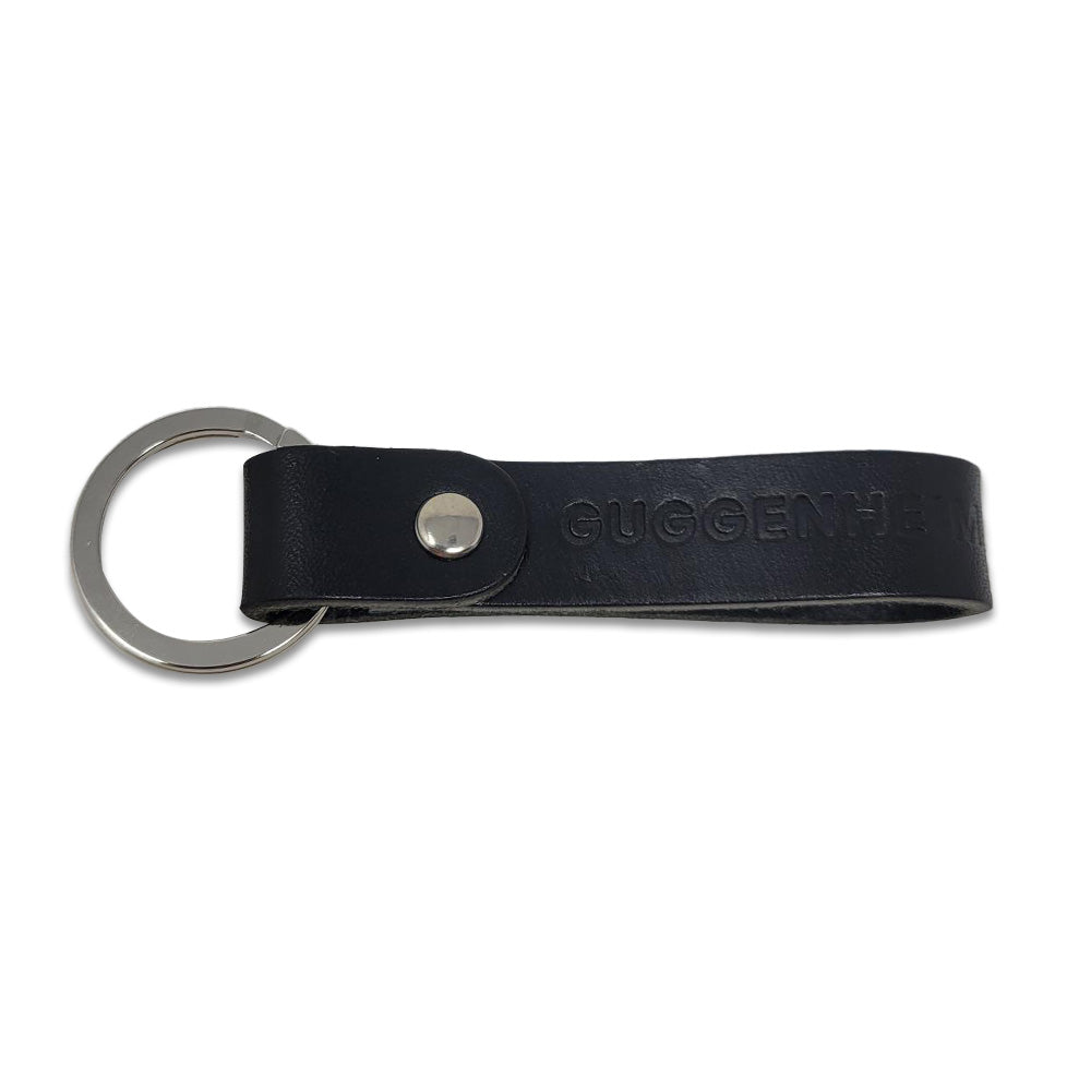 The Guggenheim Leather Keychain is crafted from black leather with a silver metal keyring and rivet. “GUGGENHEIM” is embossed on the durable leather, combining elegance with lasting style.