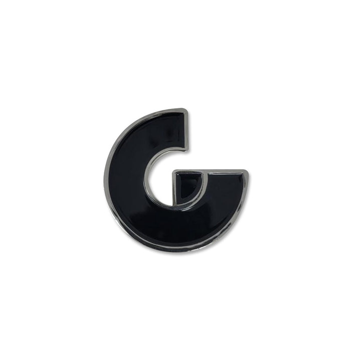 A shiny black Guggenheim Magnet, shaped as a stylized letter G with smooth, rounded edges and a metallic outline, evokes neodymium strength against a plain white background.