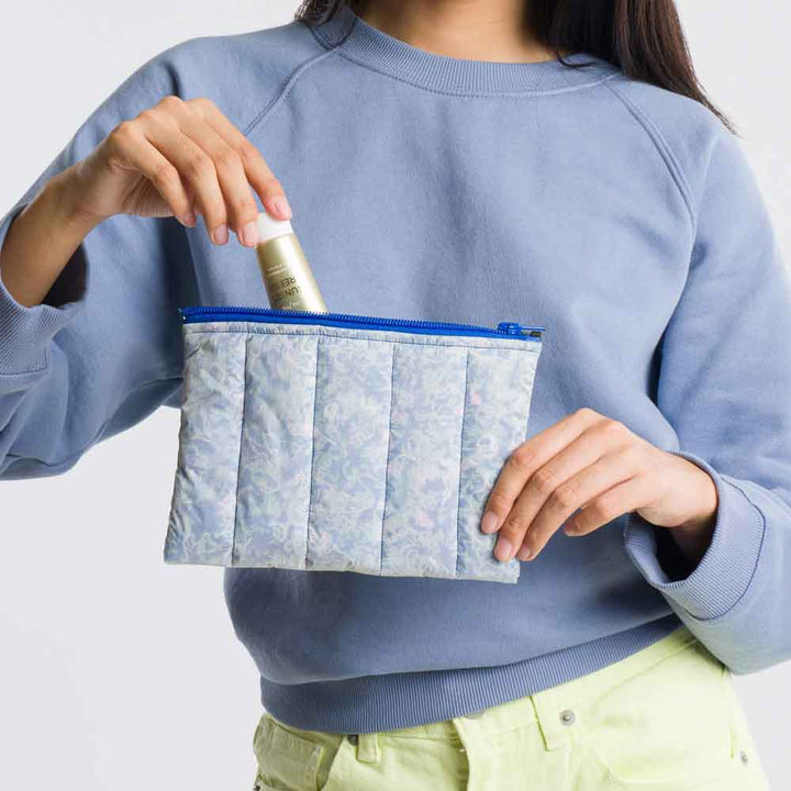 A person in a light blue sweatshirt and pale yellow pants puts a small bottle into an eco-friendly, floral-patterned Scrap Stuffed Pouch with quilting.