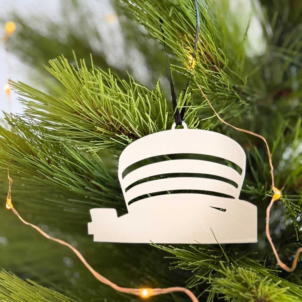 The Guggenheim Ornament, a small metal piece shaped like the iconic museum, hangs from a green Christmas tree branch. Compact enough to fit with a business card or in your wallet, it sparkles as string lights glow nearby.