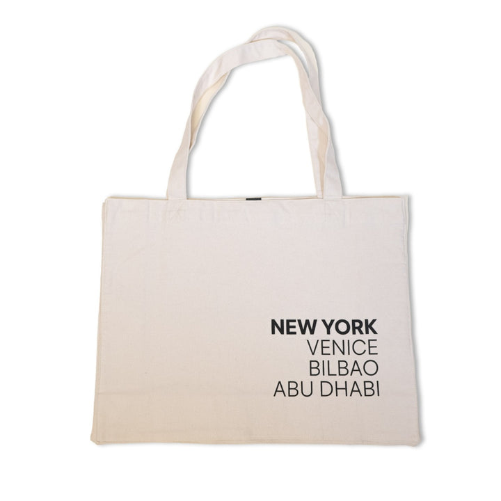 The Guggenheim Constellation Tote Bag New York, Natural is a beige organic cotton bag with two handles, featuring NEW YORK, VENICE, BILBAO, and ABU DHABI in bold black text on one side. Displayed against a white background.