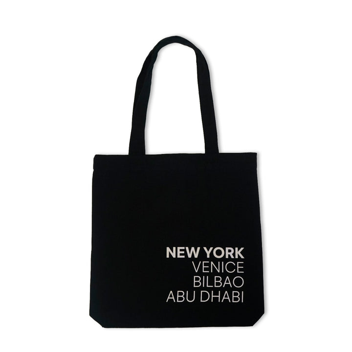 The Guggenheim Constellation Tote Bag New York, Black, is crafted from organic cotton and displays NEW YORK VENICE BILBAO ABU DHABI in white capitals—an elegant accessory for art enthusiasts.