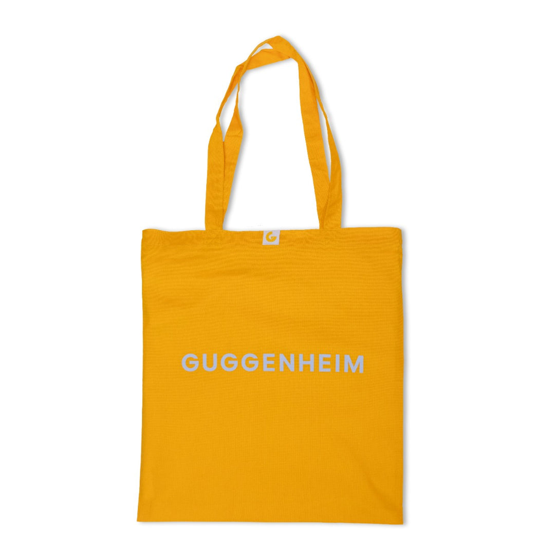 The Guggenheim Constellation Tote Bag New York in yellow is made from organic cotton, has two handles, and features "GUGGENHEIM" in white capitals on the front. The tote is shown against a plain white background.