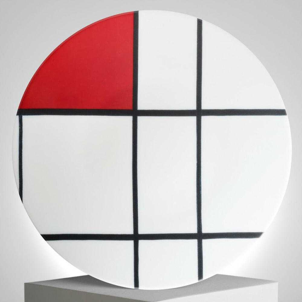 The Mondrian Composition B With Red Porcelain Plate is a round white porcelain plate featuring black grid lines and a solid red square in the upper left, inspired by Piet Mondrian’s geometric art, displayed on a gray pedestal.