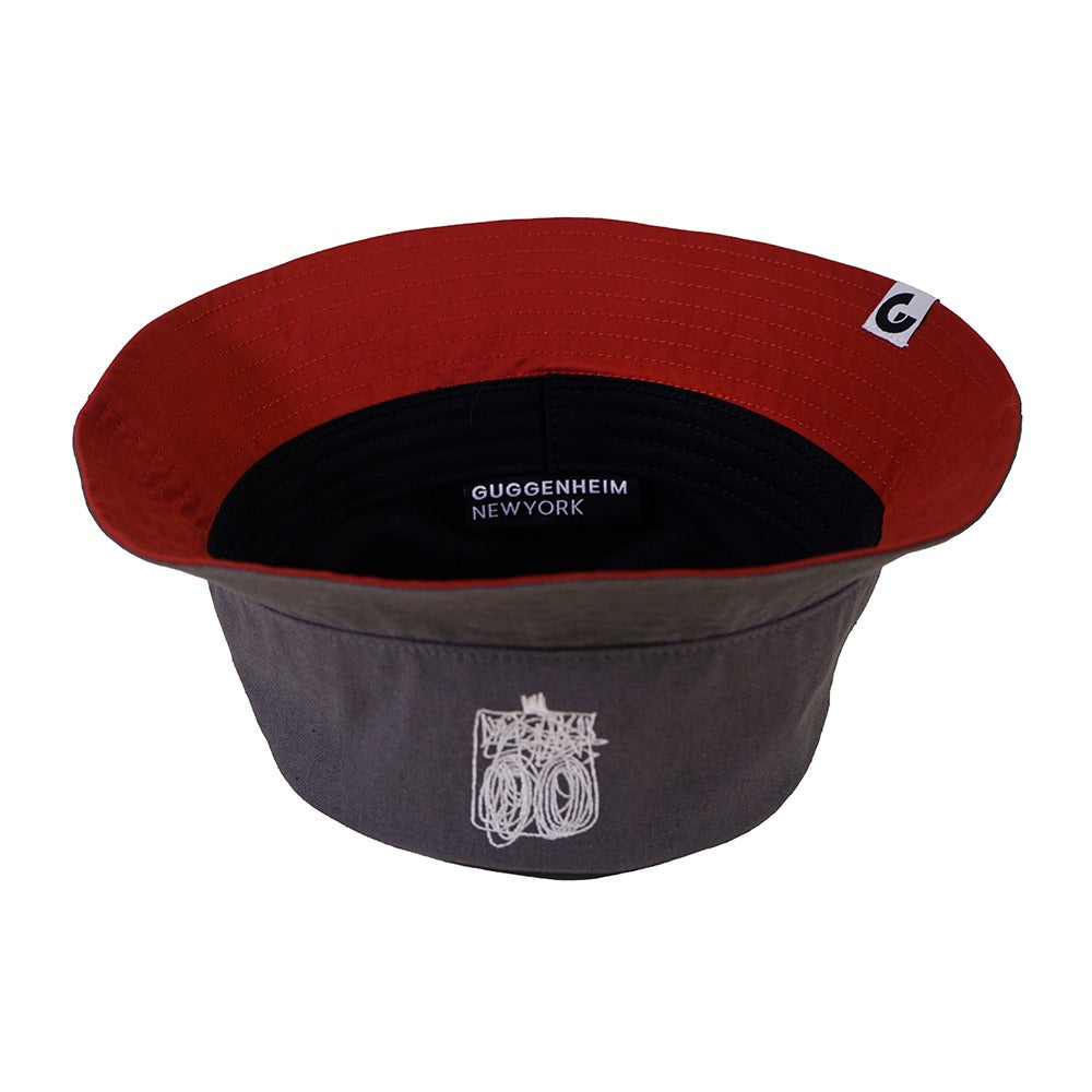 A gray unisex Rashid Johnson Bucket Cap, Untitled Anxious Men, with a wide red inner brim, white embroidered front design inspired by Anxious Men, and a Guggenheim New York label inside.