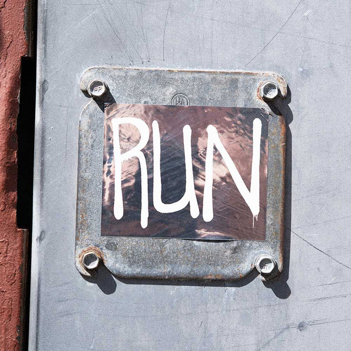 A Rashid Johnson Run Sticker, featuring the word RUN in large, white, hand-painted letters, is displayed on a 4 x 2.6 inch metal plate mounted on a wall.