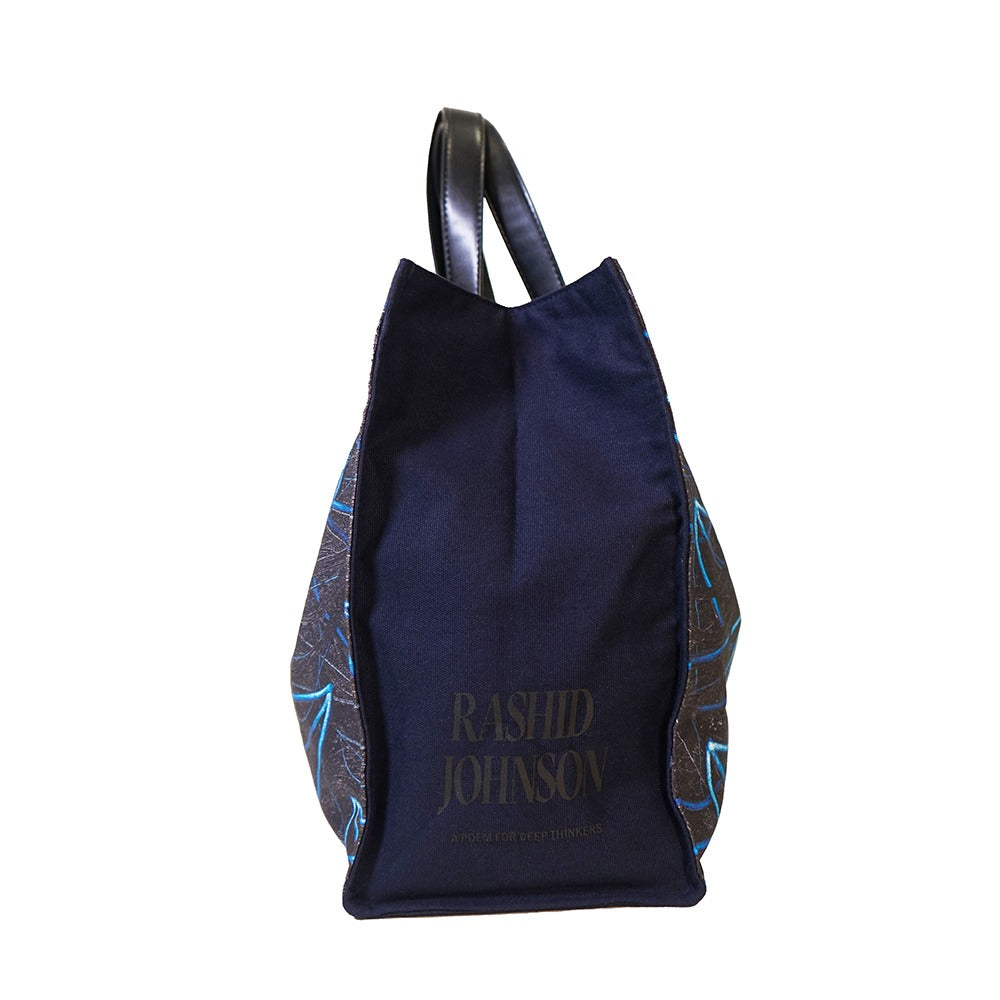 The Rashid Johnson Bag, Untitled Seascape Drawing, is a dark blue cotton canvas tote with black handles, gold artist signature on the front, and blue and white abstract patterned side panels.