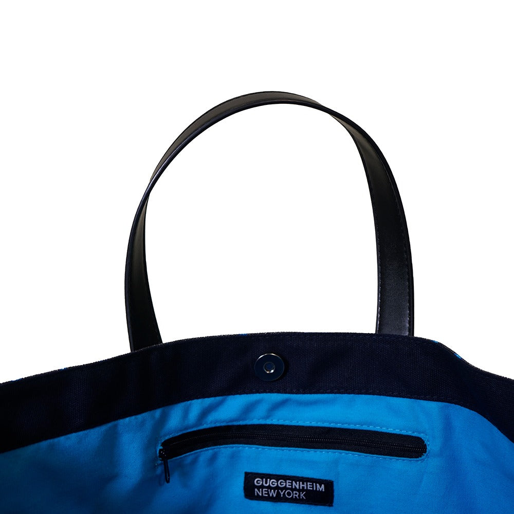 A close-up of the Rashid Johnson Bag, Untitled Seascape Drawing—an open blue and black cotton canvas bag with black handles and a zippered pocket labeled Guggenheim New York, set against a white background. Inspired by contemporary artist Rashid Johnson.