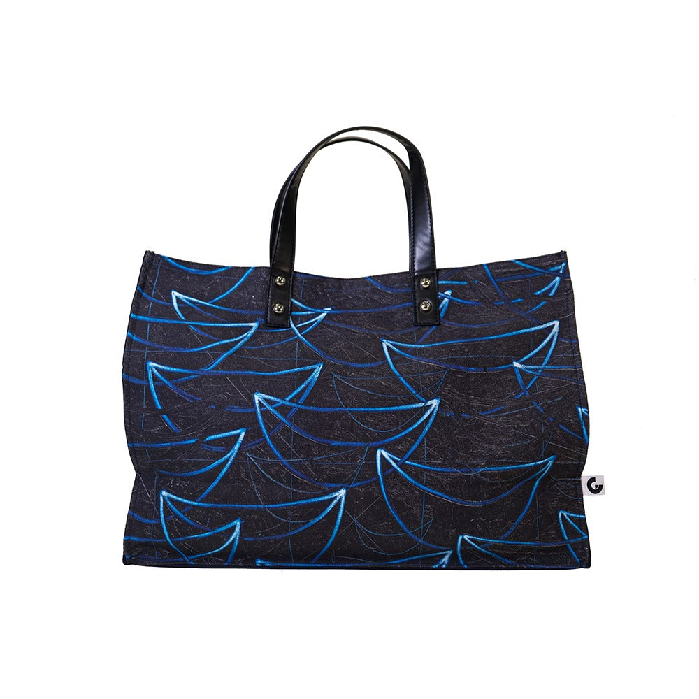 The Rashid Johnson Bag, Untitled Seascape Drawing, is a large rectangular cotton canvas bag with black leather handles and dark blue fabric featuring light blue abstract geometric patterns. A small white tag with a black logo is attached to the side.