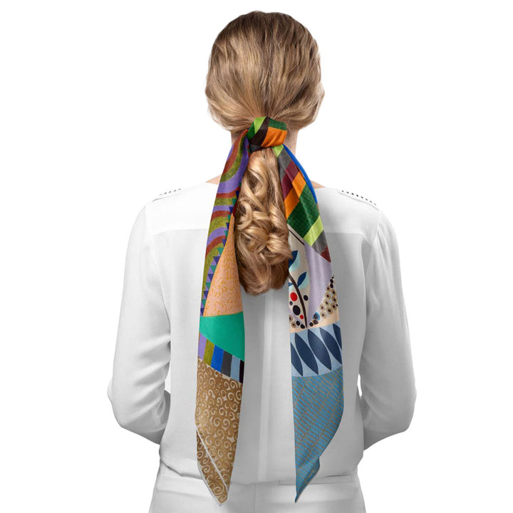 A person with long, wavy blonde hair in a white shirt ties their ponytail with the Guggenheim Beatriz Milhazes Silk Scarf, Mistura Sagrada, which features vibrant abstract art-inspired designs in green, orange, blue, and more.