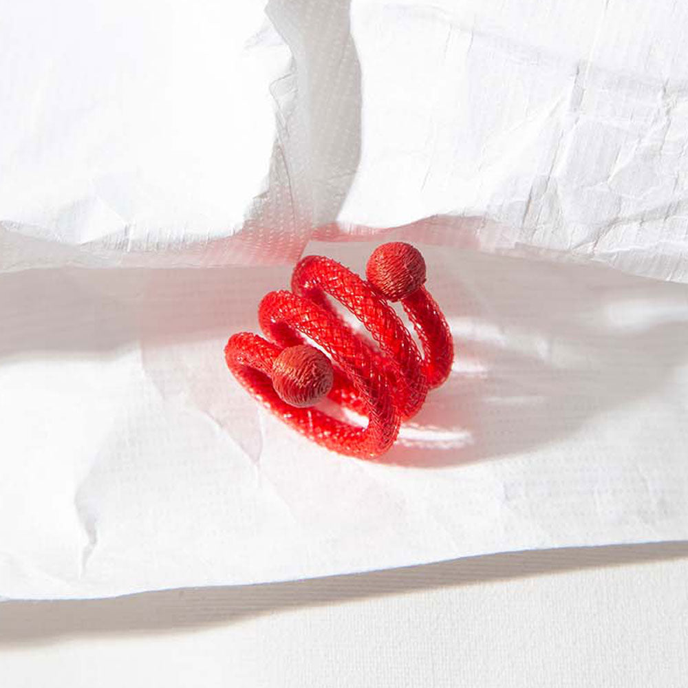 The Spiral Mesh Ring - Red, featuring a bright red spiral design with textured spheres on each end, is shown on crumpled white paper under gentle light to emphasize its handcrafted jewelry artistry.