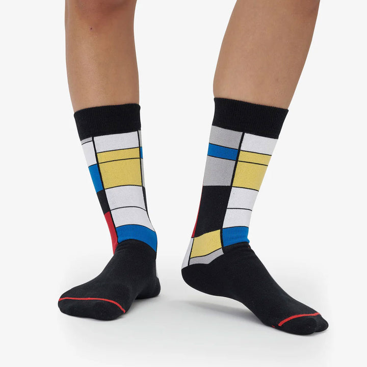 Wearing Mondrian Composition Socks, featuring bold blue, yellow, red, white, and black geometric patterns inspired by Piet Mondrian’s art. These artistic mid-calf socks stand out against a plain background.