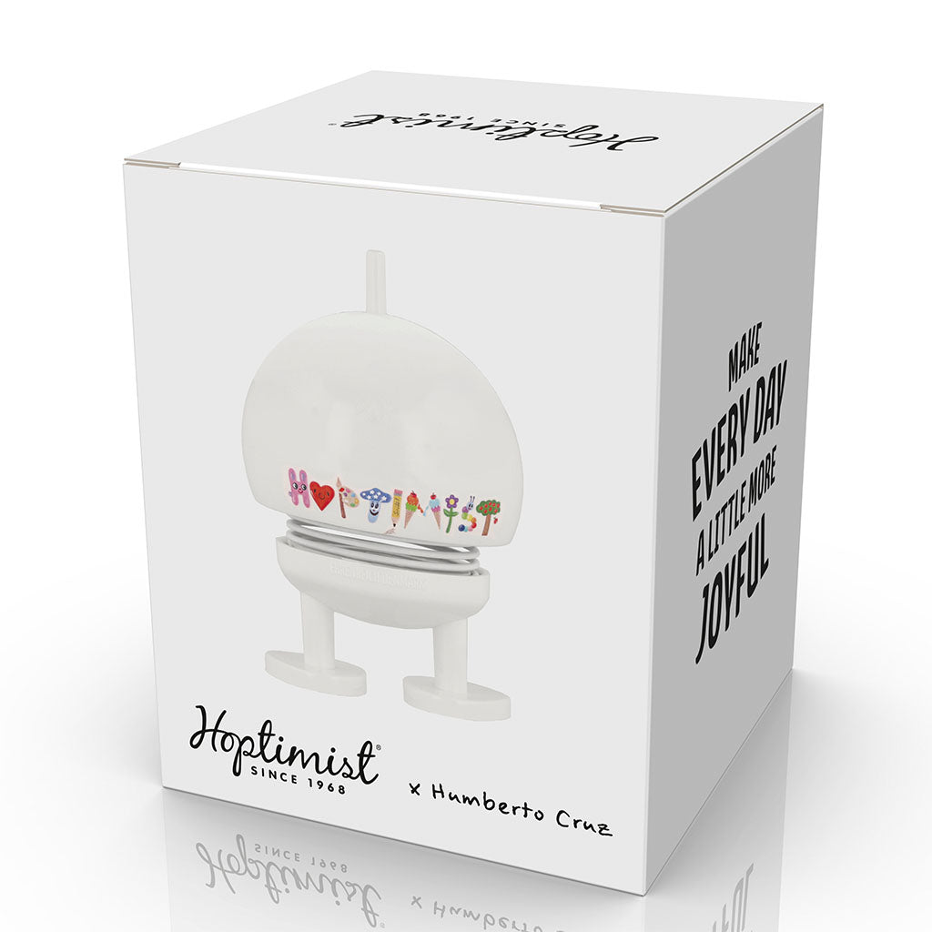A white box displays the Hoptimist x Humberto Cruz Happy Icons Figure—a round, cheerful figurine with colorful letters—plus the text: "Hoptimist since 1968 x Humberto Cruz" and "Make every day a little more joyful" in bold.