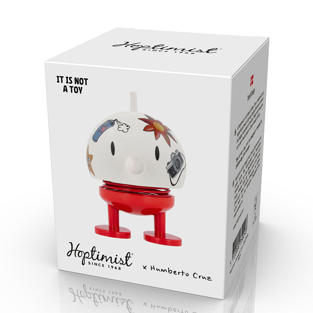 A white box displays the Hoptimist x Humberto Cruz Art is Life Medium Two-Tone Figure—a custom piece with red legs and a round head featuring Humberto Cruz art. The box reads "IT IS NOT A TOY" and includes product details and branding.