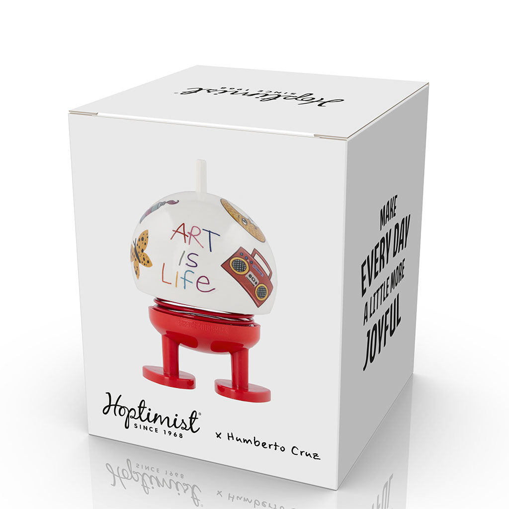 A white box displays the Hoptimist x Humberto Cruz Art is Life, Medium Two-Tone Figure—a colorful, round figurine with red legs, doodles inspired by Humberto Cruz, & "ART IS LIFE" on it. The box reads: MAKE EVERY DAY A LITTLE MORE JOYFUL.