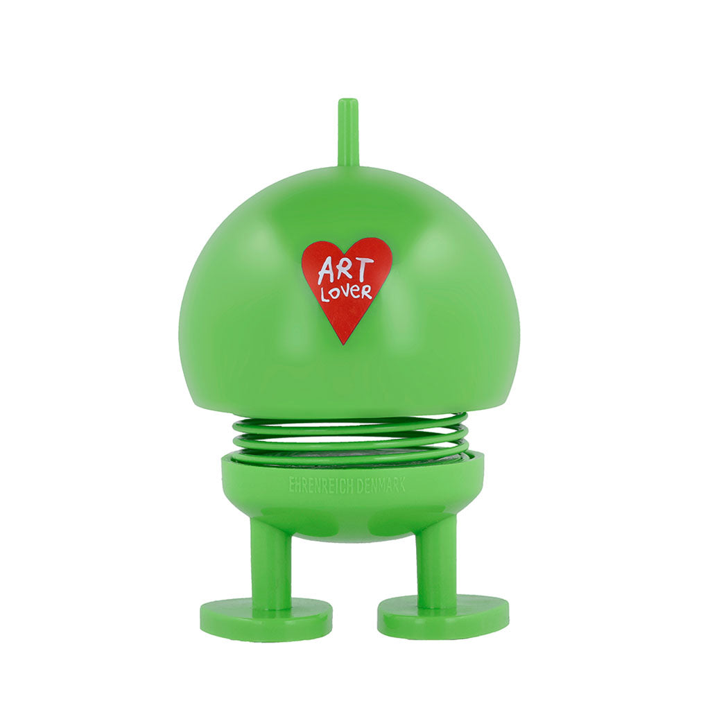 The Hoptimist x Humberto Cruz Art Lover, Green Figure is a playful, round, green figurine with spring legs, feet, a top stem, and features a red heart labeled ART LOVER on its body. Inspired by Gustav Ehrenreich’s original design.