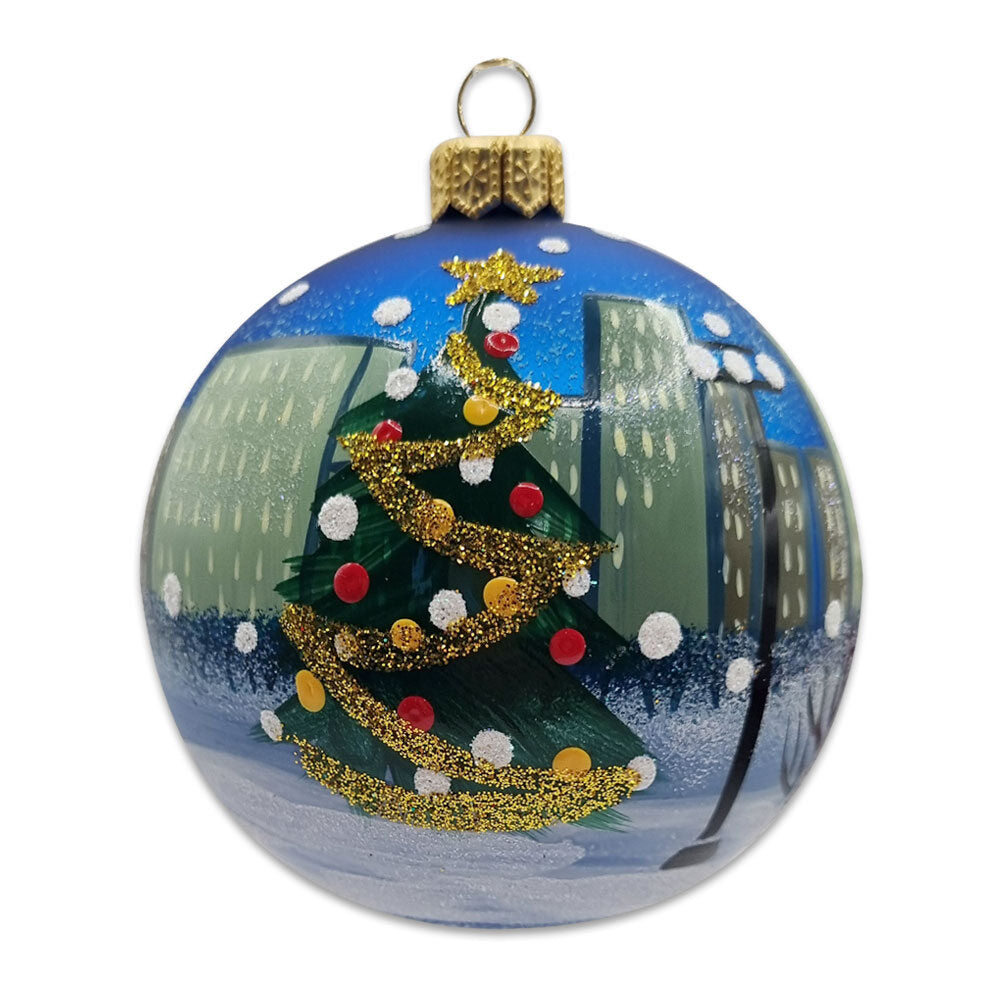 The NYC Tree Ornament is a round, mouth-blown glass Christmas ornament featuring a green tree with gold glitter garland and colorful dots against a snowy cityscape—beautifully gift-boxed for the perfect holiday surprise.
