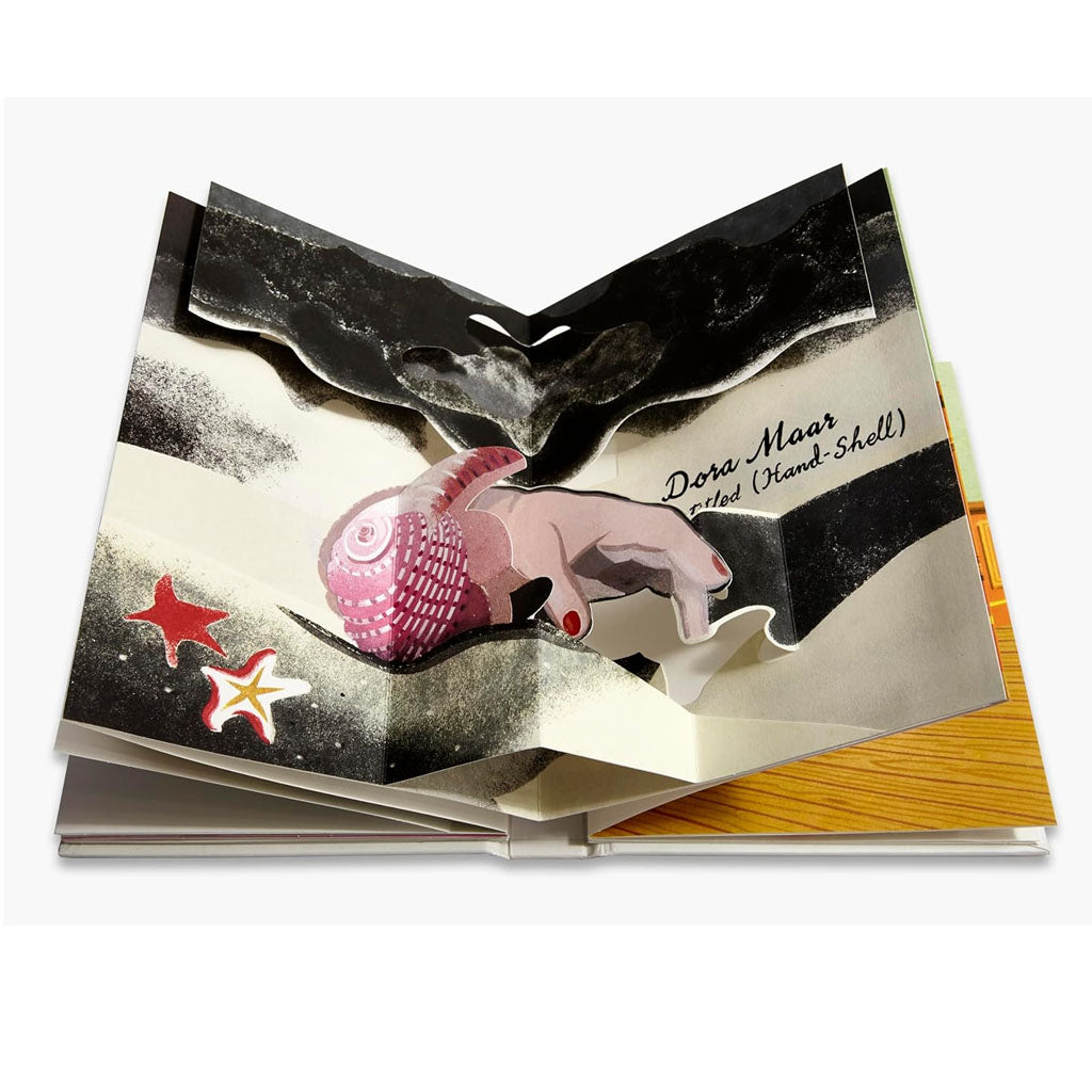 An open Pop-Up Surrealism book by Gérard Lo Monaco reveals a 3D pink seashell held by a hand, flanked by two starfish. The page reads "Dora Maar called (Hand-Shell)" in script on abstract black and white pop-up designs.