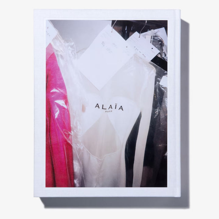 Alaïa by Seklaoui is a limited-edition book with a white cover, featuring a photo of elegant garments—including a white dress and bright pink fabric—in garment bags. The text “Alaïa Paris” honors the artistry under Pieter Mulier.