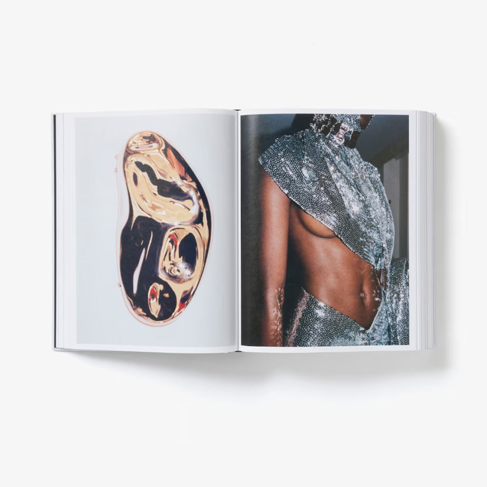 An open limited-edition book displays two pages: the left has an abstract gold and black organic shape, and the right features a close-up of someone in a sparkly, asymmetrical silver Alaïa by Seklaoui outfit revealing their midriff.