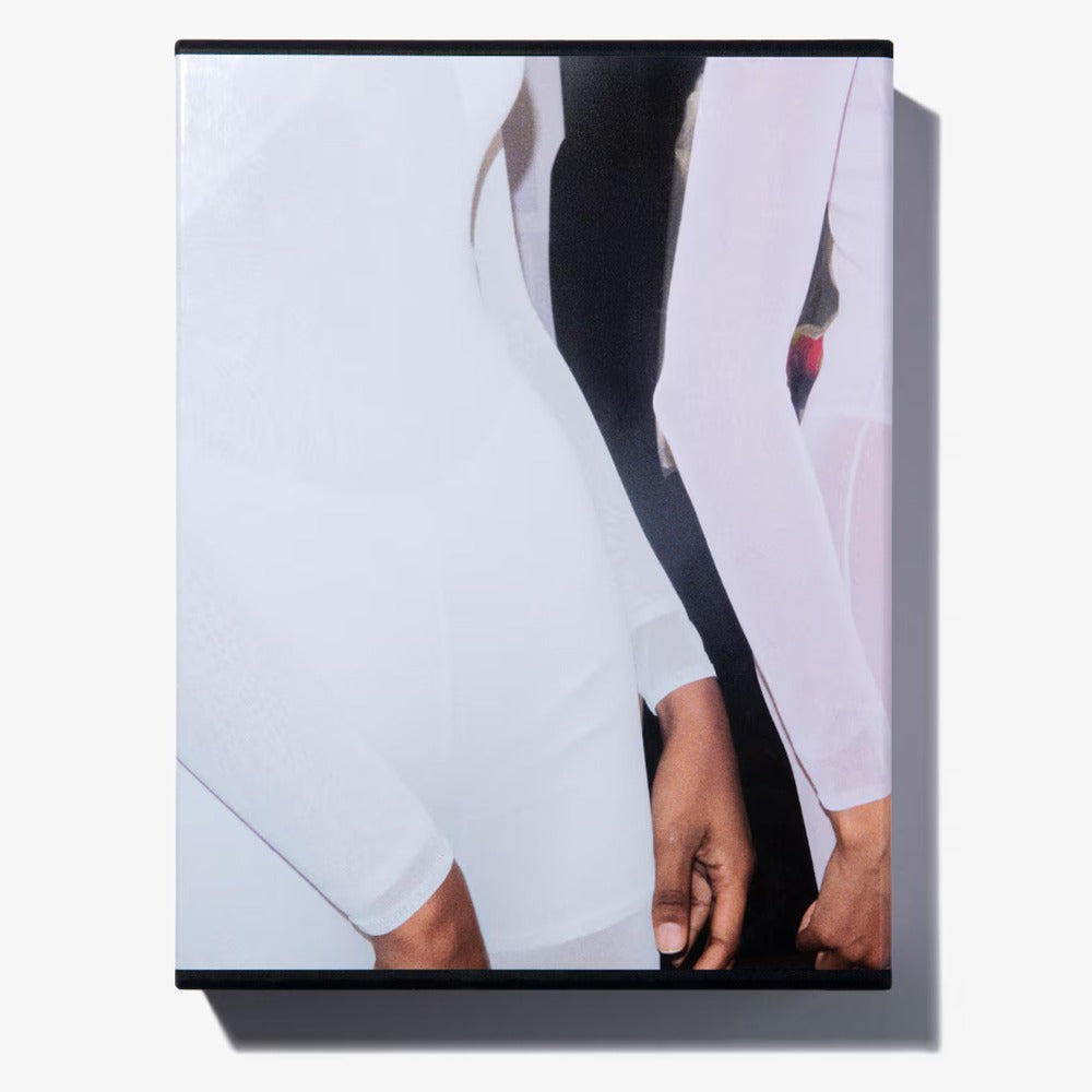 A close-up of two people side by side in fitted, long-sleeve white Alaïa by Seklaoui outfits, highlighting their torsos and arms with hands relaxed at their sides against a plain background.