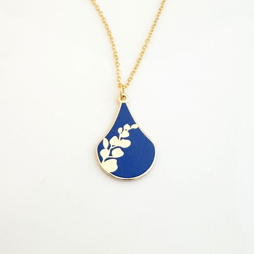 The Gabriele Münter Flower Drop Pendant Necklace features a gold-plated chain and a blue teardrop pendant with a central white floral branch design, secured with a lobster clasp and shown on a plain white background.