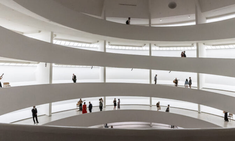 People walking along the curved ramps inside a bright, modern, white museum with multiple levels.